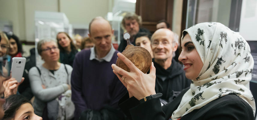 Muslim woman wearing patterned hijab (Islamic headscarf) giving a tour of the Museum, she is holding up a paper astrolabe to the audience. 